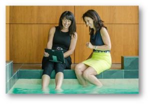 Deux femmes sont assises côte à côte au bord d'une piscine intérieure, souriant et regardant une tablette. L'une plonge ses pieds dans l'eau, tandis que l'autre s'assoit à côté d'elle, toutes deux semblant détendues et engagées.